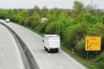 
Rückansicht eines weißen Lkw, der auf einer vierspurigen Bundesstraße an einem gelben Wegweiser vorbeifährt